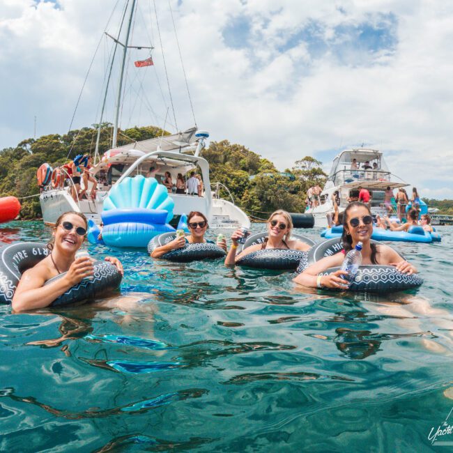 Four women in sunglasses float on inner tubes in the water, holding drinks and smiling. Behind them are two boats with more people relaxing and socializing, set against a backdrop of trees and a partly cloudy sky.