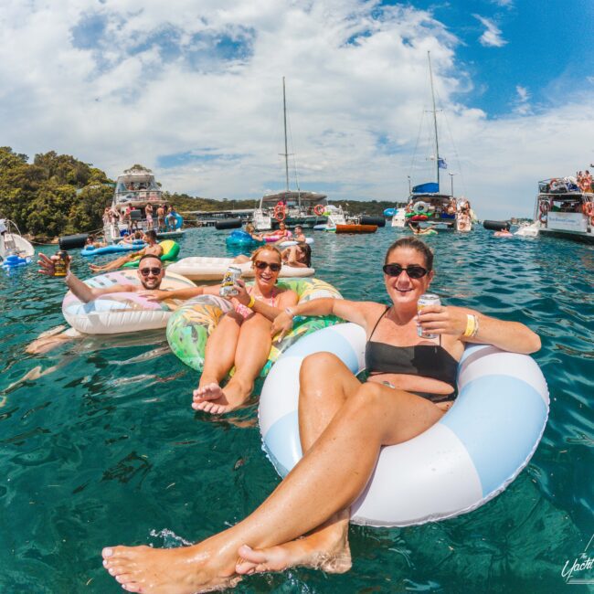 A group of people relax on inflatable tubes in clear blue water, smiling and holding drinks, with boats and other people in the background under a sunny sky.
