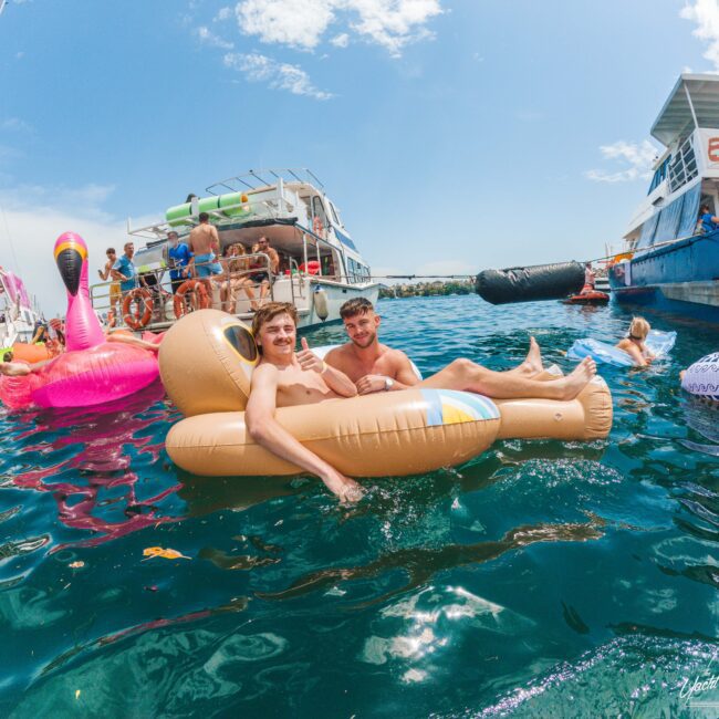 Two men smiling and relaxing on a large inflatable sloth in blue water, surrounded by other people, colorful floaties, and boats on a sunny day.
