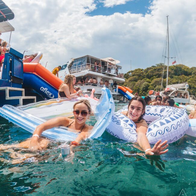 Two women smile and relax on inflatable pool floats in clear blue water near a docked boat, with other people and boats in the background under a partly cloudy sky.