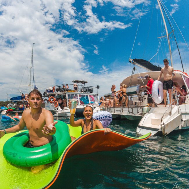 A group of people enjoy a sunny day on the water, relaxing on pool floats and boats. Two people float in the foreground, smiling and holding drinks, while others gather on boats in the background under a partly cloudy sky.