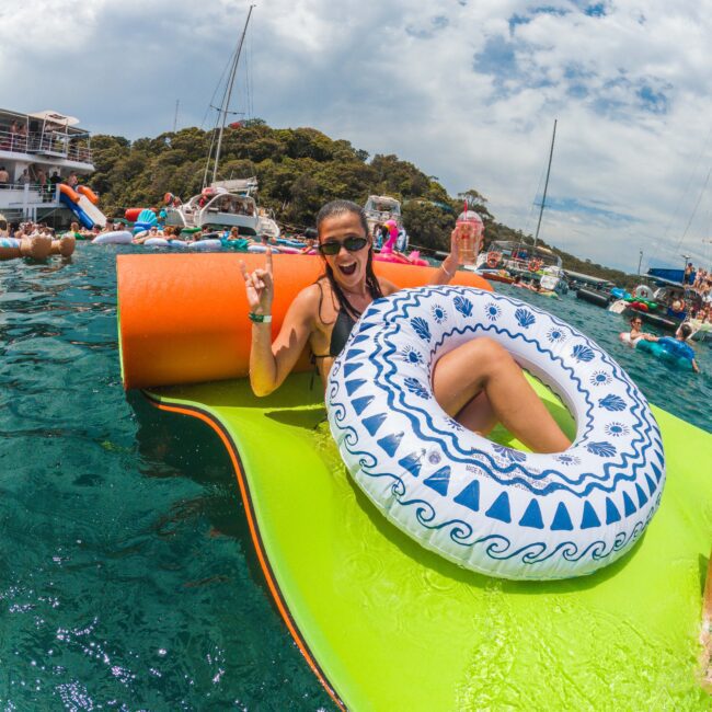 A woman in sunglasses smiles and makes a peace sign while sitting on a green float with an inflatable ring on a lively, sunny day on the water surrounded by boats and other people.