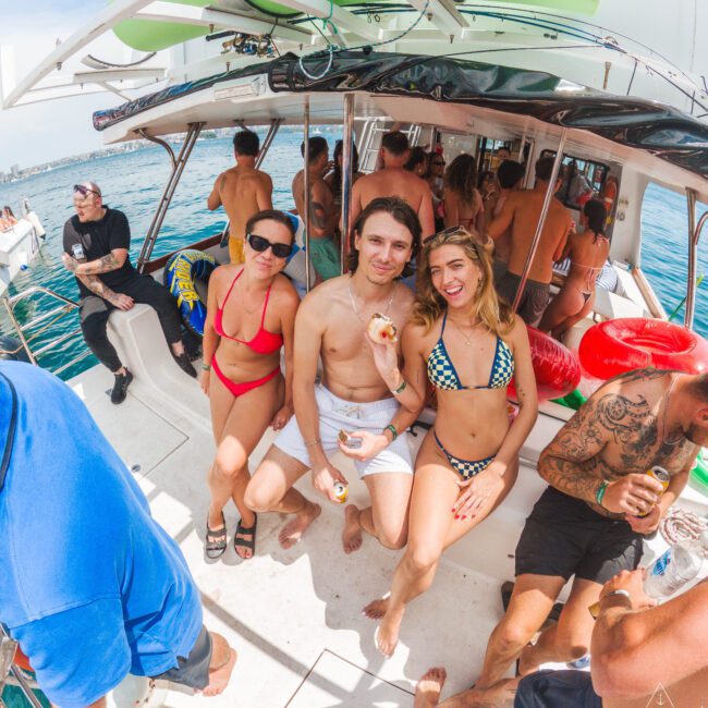 A group of people in swimsuits relax and socialize on a boat in sunny weather. Three people sit together smiling at the camera, while others mingle around them. The sea and coastline are visible in the background.