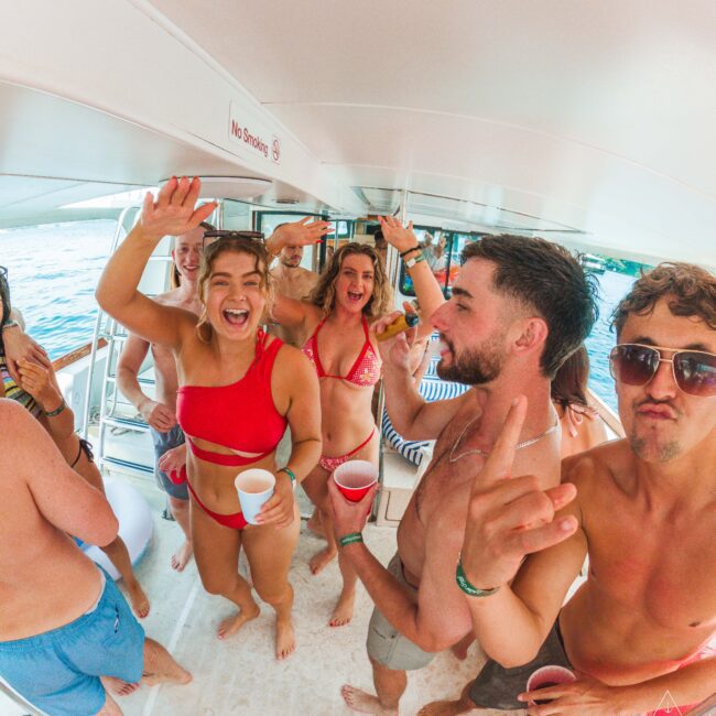 A group of young adults in swimsuits laugh and dance on a boat, holding red cups. The boat is on the water, and the atmosphere is lively and carefree. A "No Smoking" sign is visible overhead.