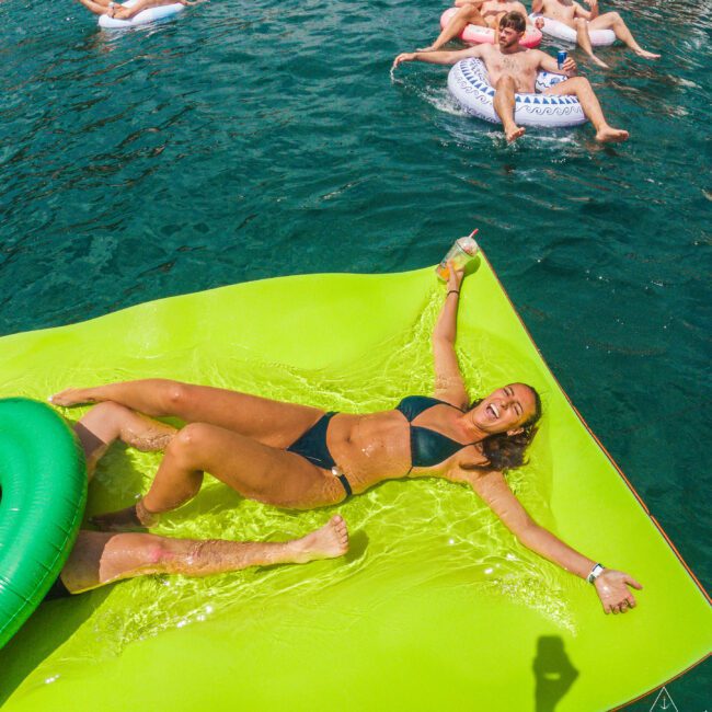 A woman in a black bikini relaxes on a large green float in the water, smiling and holding a drink. Other people are lounging on inflatables in the background, enjoying a sunny day.
