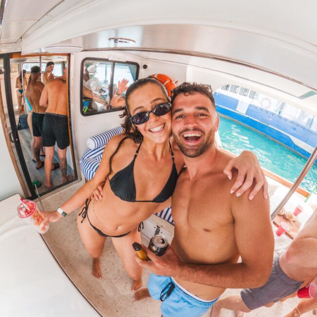 A smiling woman in a black bikini and sunglasses and a man in swim trunks pose for a selfie on a boat. Other people are in the background, and turquoise water is visible outside. Both are holding drinks and appear happy.