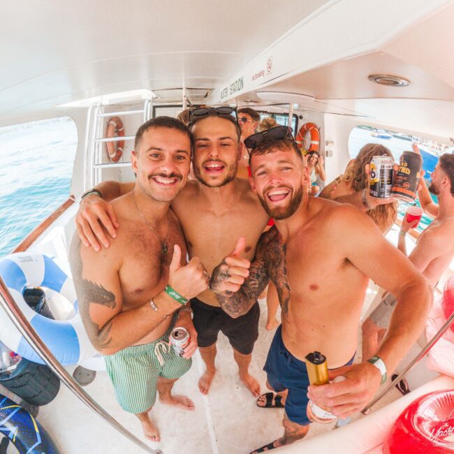 Three shirtless men smiling with their arms around each other, giving thumbs up and holding drinks on a boat with other people in the background, enjoying a sunny day on the water.