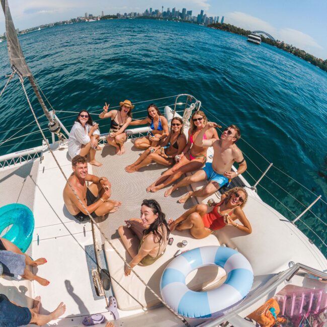 A group of twelve people in swimwear relax, smile, and pose for the camera on the deck of a boat, with blue water and a city skyline in the background under a partly cloudy sky.
