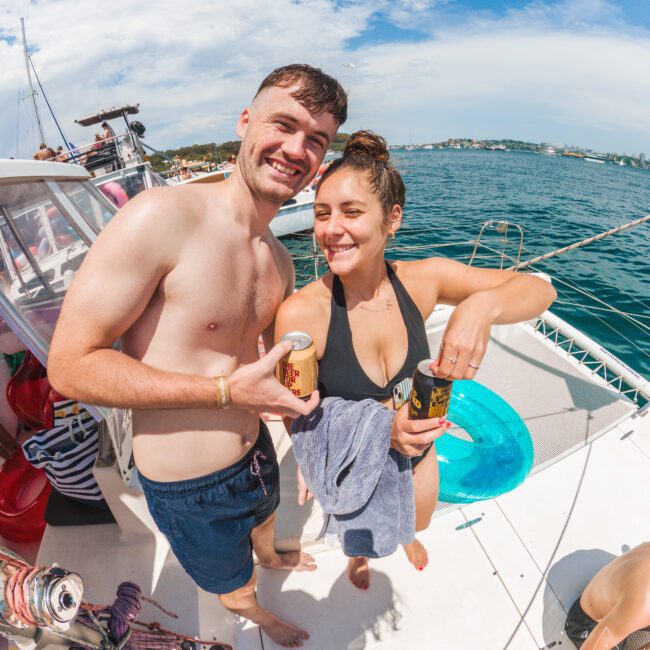 A smiling man and woman in swimsuits hold canned drinks on a boat, with blue water and a clear sky in the background. The woman has a towel around her waist, and other people and boat equipment are visible nearby.