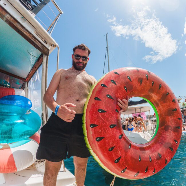 A man in swim trunks stands on a boat holding a large, round watermelon-shaped inflatable. He is smiling, wearing sunglasses, and there are people and other colorful inflatables in the background on a sunny day.