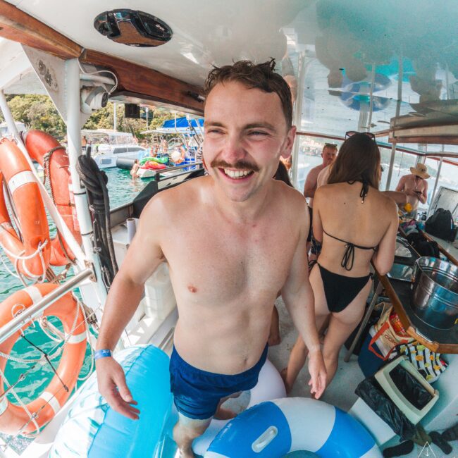 A smiling man in swim trunks and a mustache stands on a boat holding a blue and white inner tube, surrounded by other people in swimwear. The boat is docked on clear blue water with trees visible in the background.