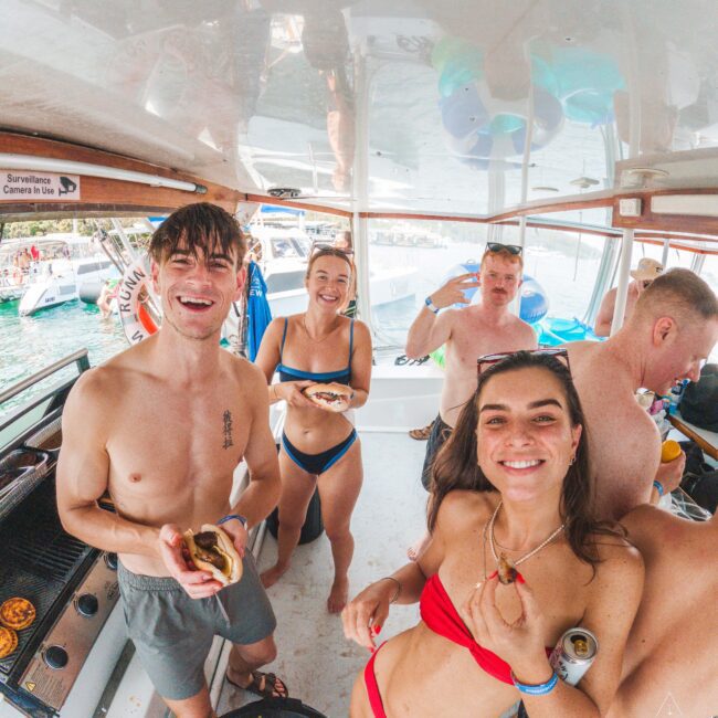 Five young adults in swimwear smile and eat food on a boat. The group appears to be enjoying a party with other boats and people visible on the water outside. The scene is lively and casual.