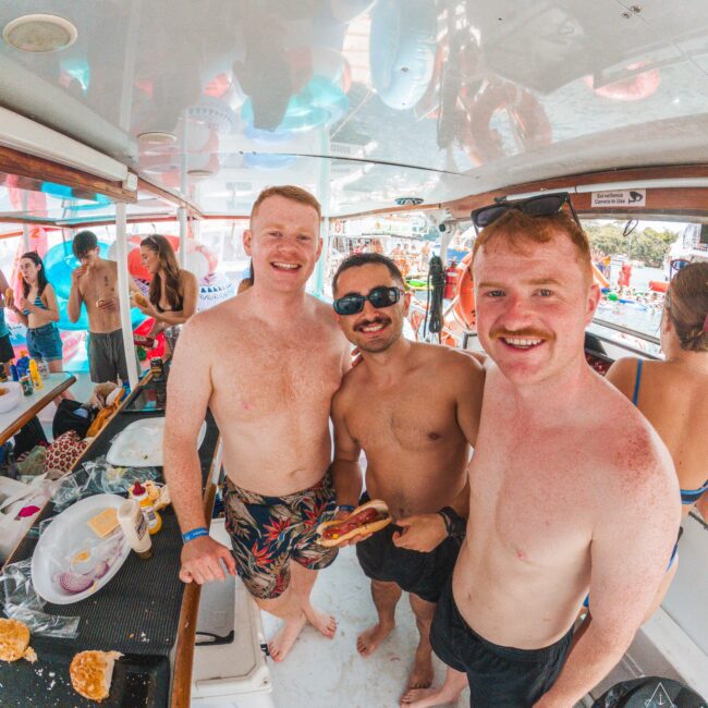 Three smiling men in swim trunks stand on a boat, one holding a sandwich. Other people and food are visible in the background, indicating a lively gathering or party on the water.