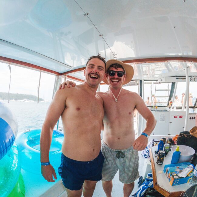 Two smiling men in swim trunks stand arm-in-arm on a boat, wearing sunglasses and wristbands. Inflatable pool floats and snacks are visible, with a sunny waterfront and other people in the background.