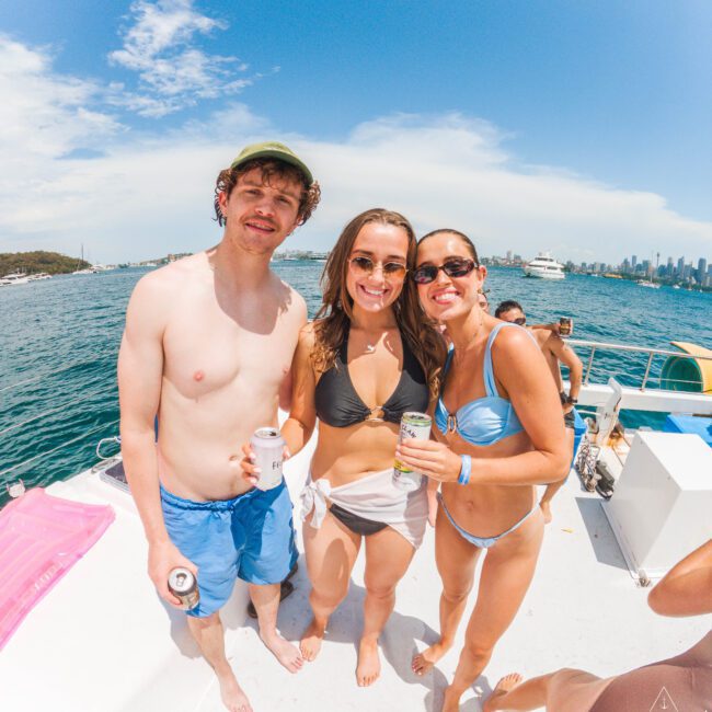 Three people in swimsuits smiling and holding drinks on a boat, with blue water and city skyline in the background on a sunny day.
