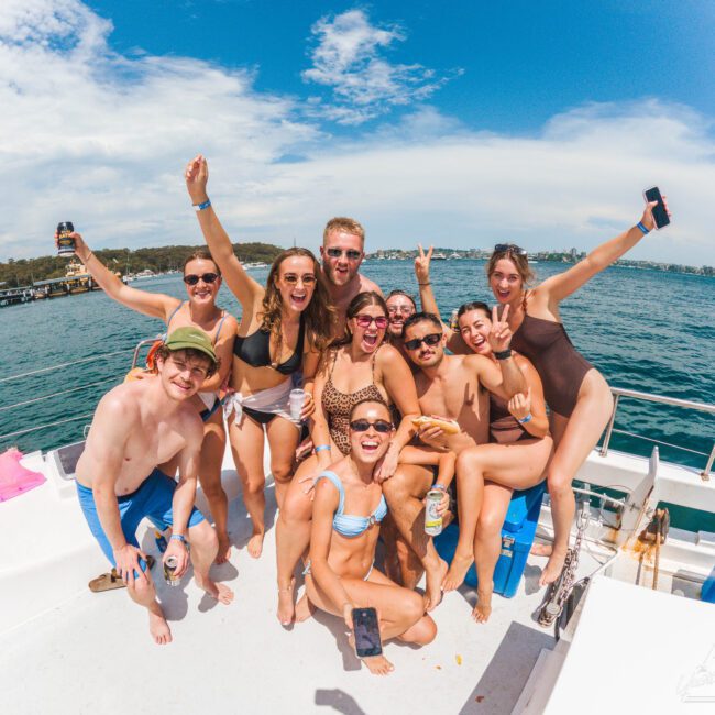 A group of young adults in swimsuits smiling, laughing, and posing together on a boat under a sunny sky, with blue water and a distant shoreline in the background.
