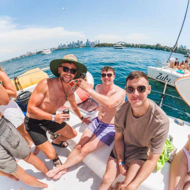 Three men sit and smile on a boat under sunny skies, with city buildings and other boats in the background. One man wears a large hat and sunglasses; all appear relaxed, enjoying a day out on the water.