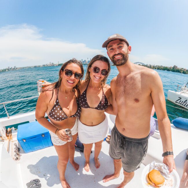 Three smiling people in swimwear stand together on a boat under a sunny sky. One woman holds a drink, the man holds a plate with a burger, and the ocean and city skyline are visible in the background.