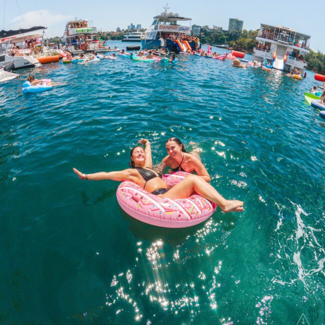 Two women smile and relax together on a pink donut-shaped pool float in clear blue water, surrounded by boats and other people enjoying a sunny day on the water.