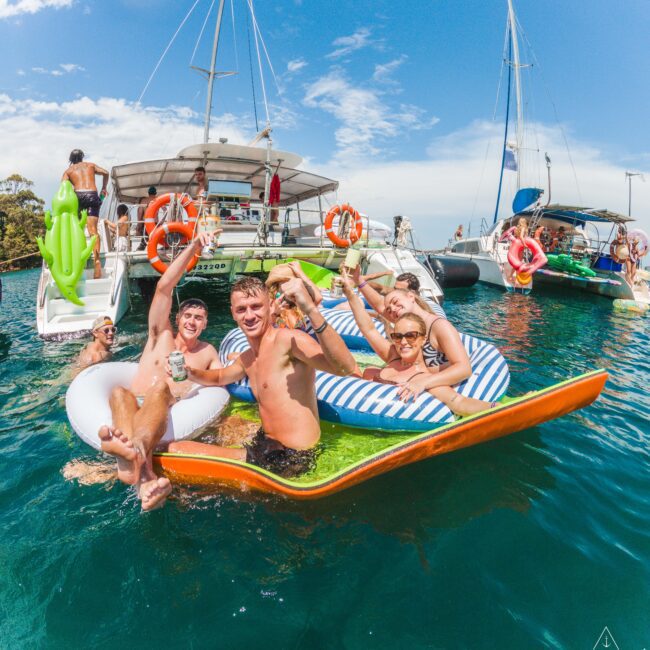 A group of smiling young adults relax on inflatable floats in the water near yachts, holding drinks and making peace signs, with more people socializing on the boats under a sunny sky.
