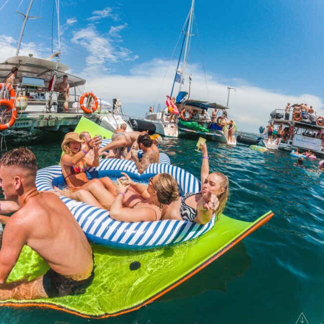 People relax and enjoy drinks on inflatable rafts in the water near several boats on a sunny day, with blue skies and vibrant, festive atmosphere.