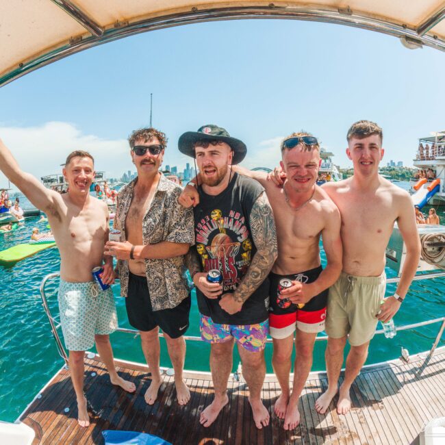 Five smiling men pose together on a boat, wearing swimsuits and holding drinks, with a lively party scene and other boats in the background on a sunny day by the water.