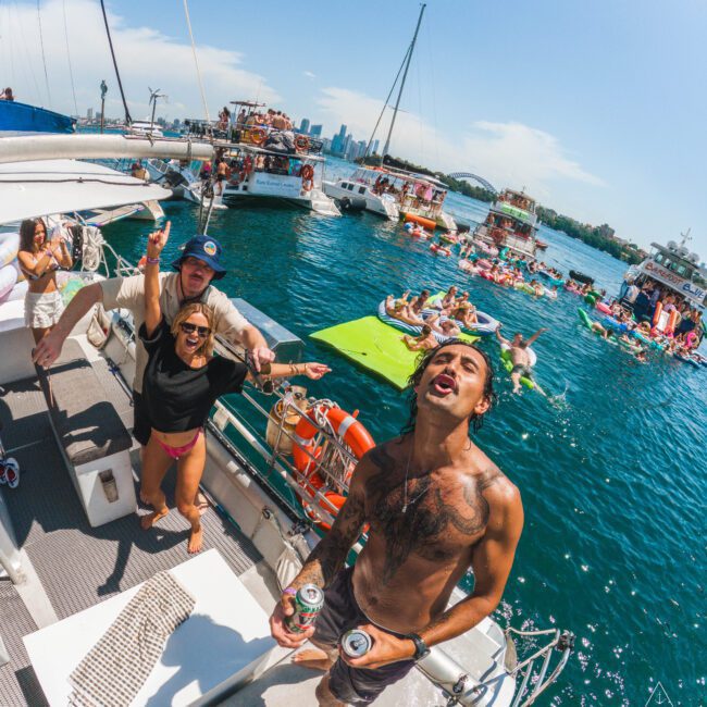 Young adults enjoy a lively boat party on a sunny day, with one shirtless man in the foreground holding drinks and people dancing behind him. Other boats and swimmers are visible on the blue water.
