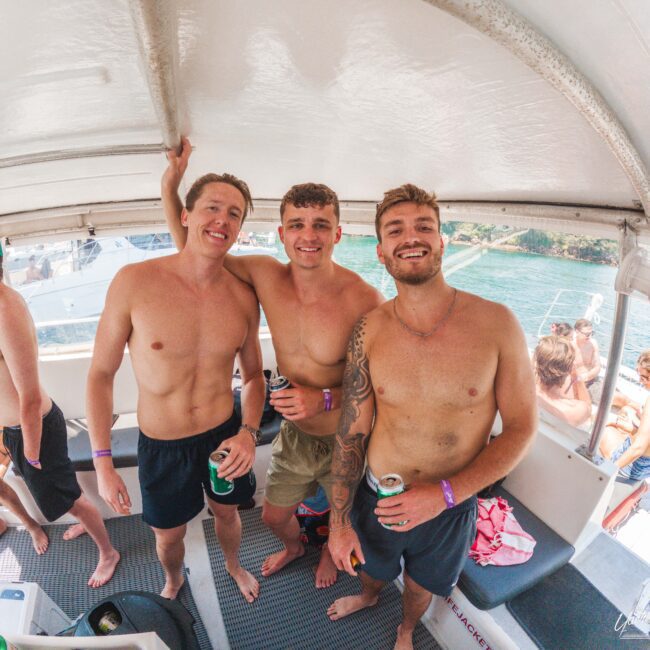 Three smiling men in swim trunks stand close together on a boat, each holding a drink. Other people are relaxing and socializing in the background, enjoying a sunny day near the water.