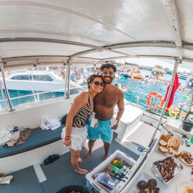 A smiling couple in swimsuits poses on a boat with food and drinks, surrounded by water and other boats in the background on a sunny day.