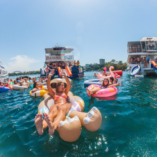 People relax on colorful inflatable pool floats in the water near boats during a sunny day. Many are wearing swimsuits, smiling, and enjoying the lively atmosphere of a boat party.