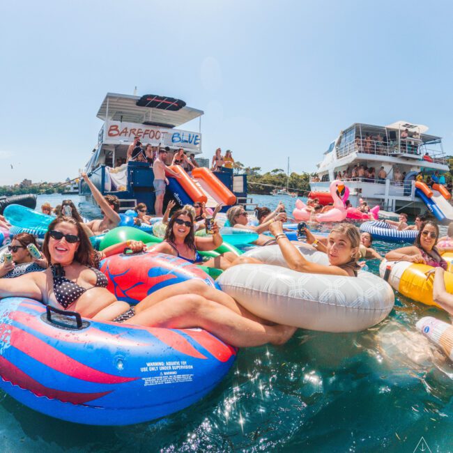 People relax on colorful pool floats in the water near boats during a sunny day. Some smile and wave, while others enjoy the festivities. The atmosphere is lively and fun, with clear blue skies overhead.