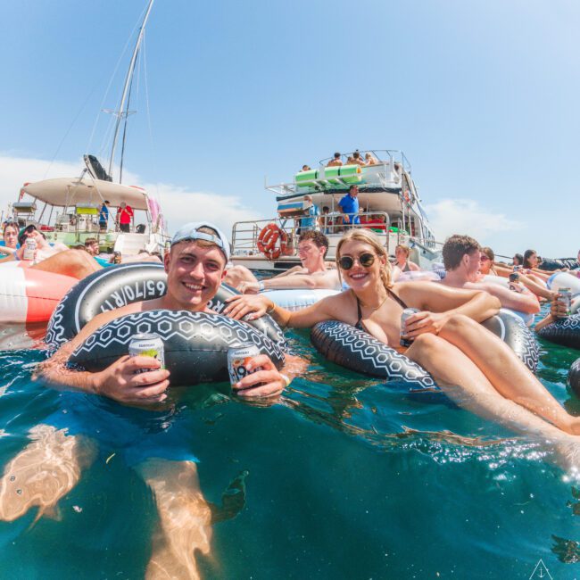 Two people smile while floating on inflatable tubes in the water, holding drinks. Behind them, others relax on tubes and a large boat under a clear, sunny sky. The atmosphere is lively and festive.
