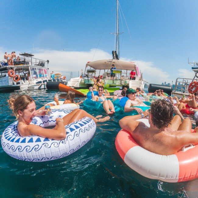 People relax on colorful inflatable tubes in the ocean near anchored boats under a sunny sky, enjoying drinks and socializing. The atmosphere is lively and festive, with clear blue water and city skyline in the background.