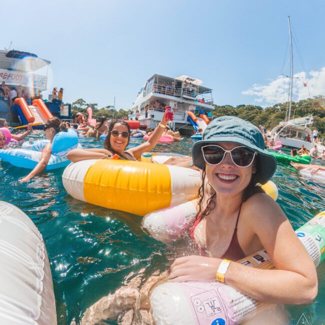 Two women wearing sunglasses and swimsuits smile while floating on colorful inflatable pool toys in sunny, blue-green water. Other people and boats are visible in the lively background.