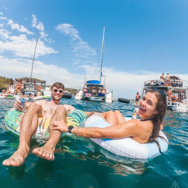 A man and woman relax on inflatable pool floats in the ocean, smiling and holding hands, with yachts and other people in the background under a bright blue sky.
