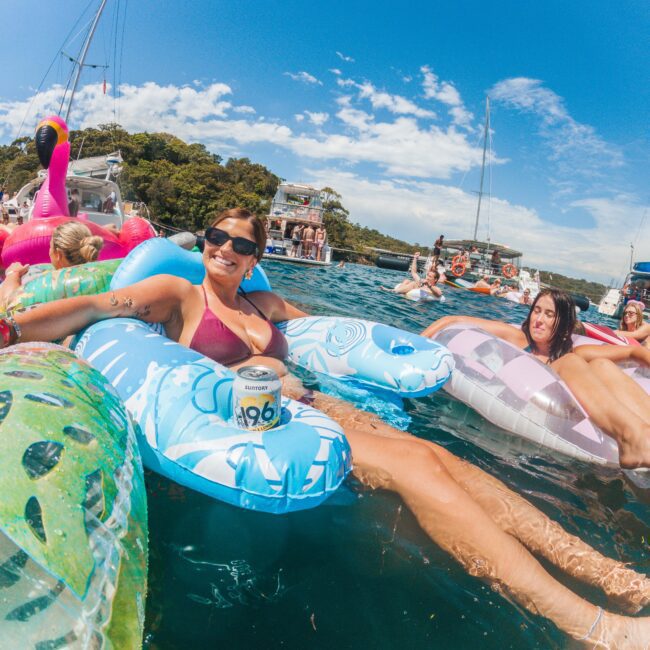 People relaxing on colorful inflatables in the water, with a woman in sunglasses and a bikini smiling at the camera and holding a drink. Boats and more people are in the background under a sunny, blue sky.