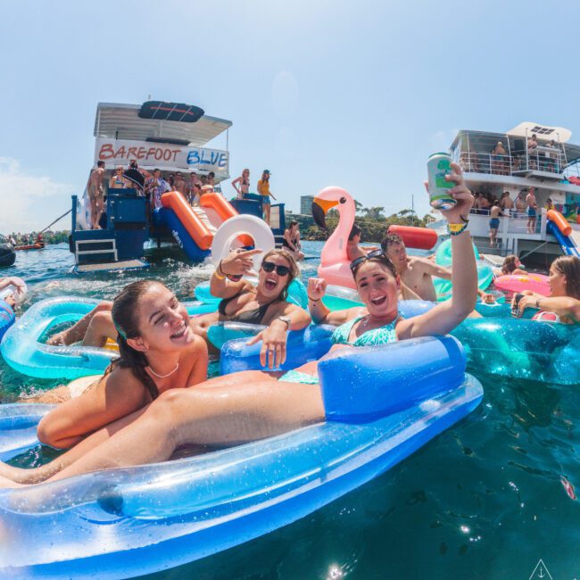 A group of young people relax and smile on inflatable pool floats in the water, holding drinks, near a floating platform with more people and a sign that reads "BAREFOOT BLUE." The weather is sunny and festive.