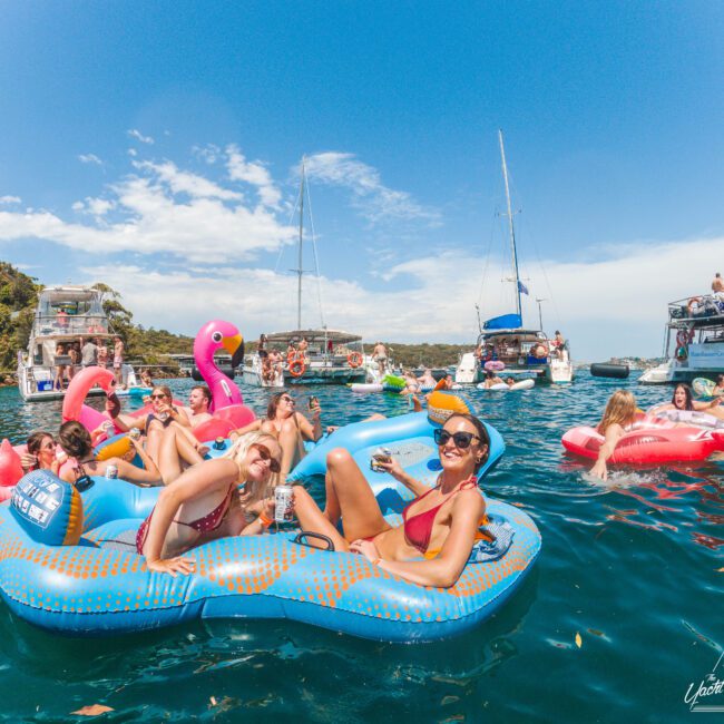 People relaxing on colorful inflatable floats in a blue, sunny bay surrounded by boats. Some are smiling and holding drinks, while others lounge on pool floats shaped like flamingos. The scene is lively and festive.