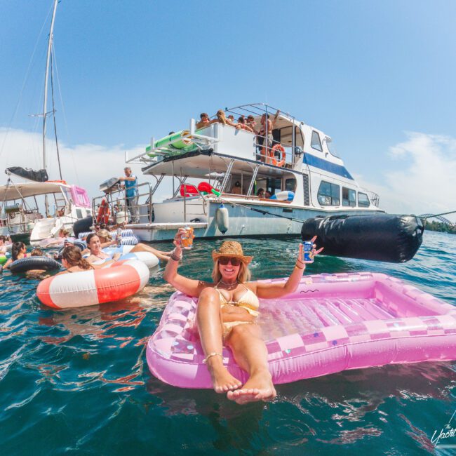 A woman in a bikini smiles and raises drinks while lounging on a pink float in the water near several others on floats, with people on a large boat in the background under a sunny sky.