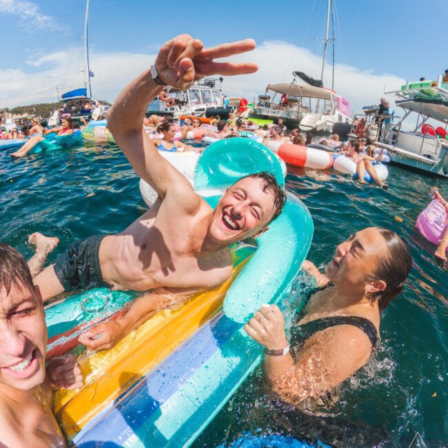 Three people smile and laugh while enjoying a pool float in the water at a busy boat party. One person flashes a peace sign. Other partygoers and boats are visible in the background under a sunny sky.