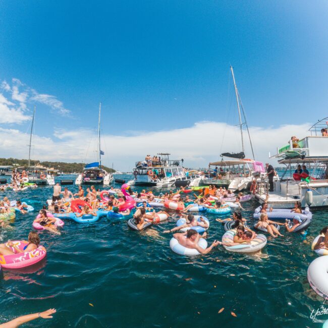 Crowd of people relaxing on colorful inflatable floats in the water near several anchored boats, under a bright blue sky. The atmosphere is lively and festive, with many enjoying the summer day.