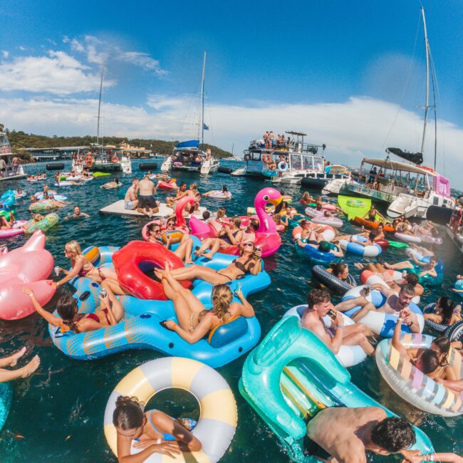 A lively scene of many people relaxing on colorful inflatable floats in the water, surrounded by boats on a sunny day. The floats are shaped like flamingos, unicorns, rings, and more, creating a festive atmosphere.