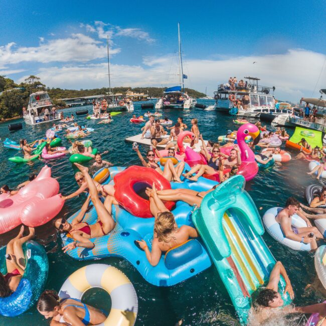 Crowd of people relaxing on colorful inflatables and pool floats in the water, surrounded by boats under a blue sky. The scene is lively and festive, with many people enjoying the sunny weather.
