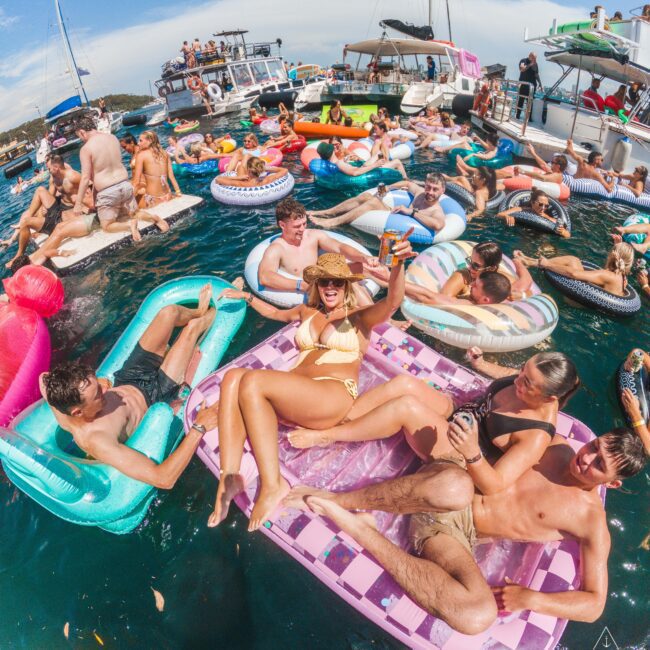A large group of people are lounging on colorful inflatables and floating in the water near boats, enjoying a sunny day. Many are smiling, raising drinks, and appear to be having a lively, festive time together.