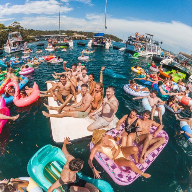 A lively group of people on colorful inflatables and rafts enjoy a sunny day on the water, surrounded by boats and others relaxing or partying on floaties. The atmosphere is festive and vibrant under a blue sky.