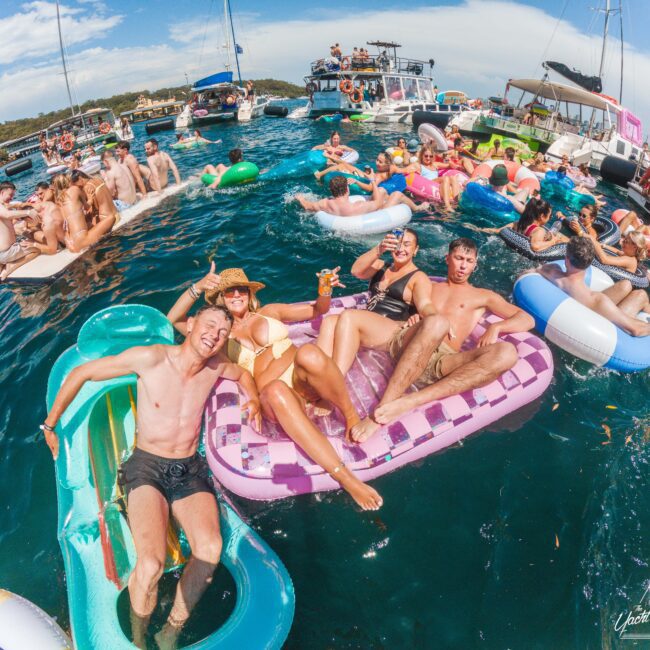 A lively group of people relax and have fun on colorful inflatables in the water near several boats, enjoying a sunny day at a floating party. Everyone looks happy and carefree.