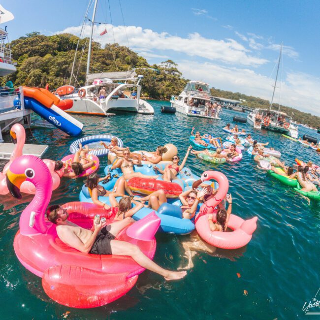 People relax on colorful inflatable pool floats in the water near several boats on a sunny day. Many floats are shaped like flamingos or other animals, and the scene is lively with groups enjoying the atmosphere.