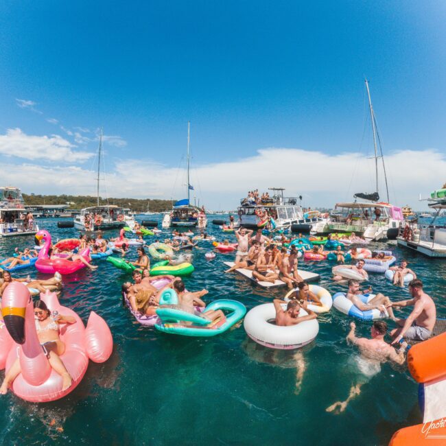 A large group of people relax on colorful inflatable floats in the water near several anchored boats on a sunny day, enjoying a lively outdoor pool party under a clear blue sky.