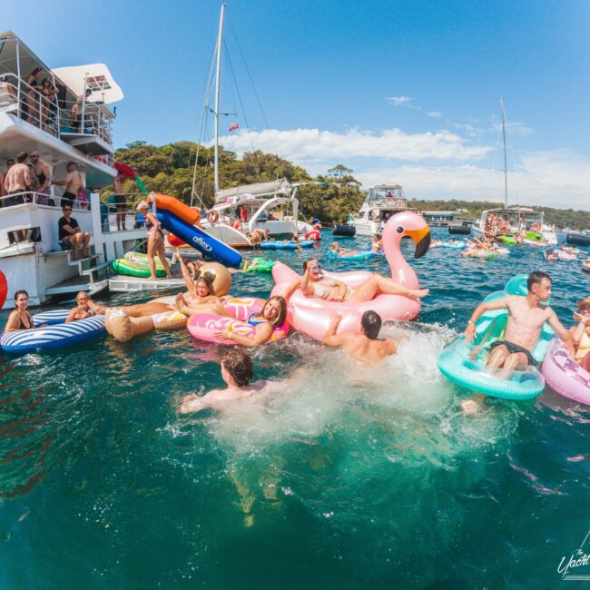 A lively group of people enjoy a party on the water with colorful inflatable pool floats and boats, swimming and relaxing under a sunny sky near a tree-lined shore.