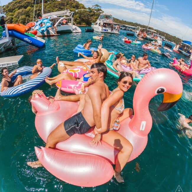 A group of people float on colorful inflatables in a sunlit bay. In the foreground, two people smile while sitting on a large pink flamingo float. Boats and others on floats are visible in the background under a blue sky.